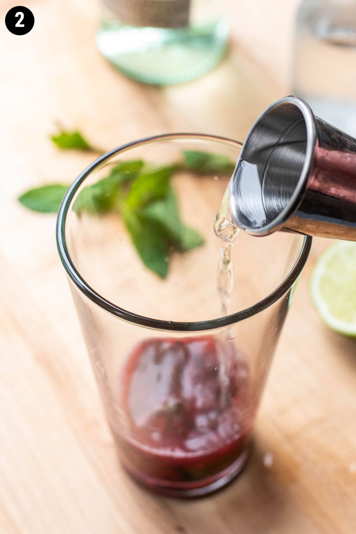 A double jigger pouring rum into a cocktail shaker glass with muddled blackberry.