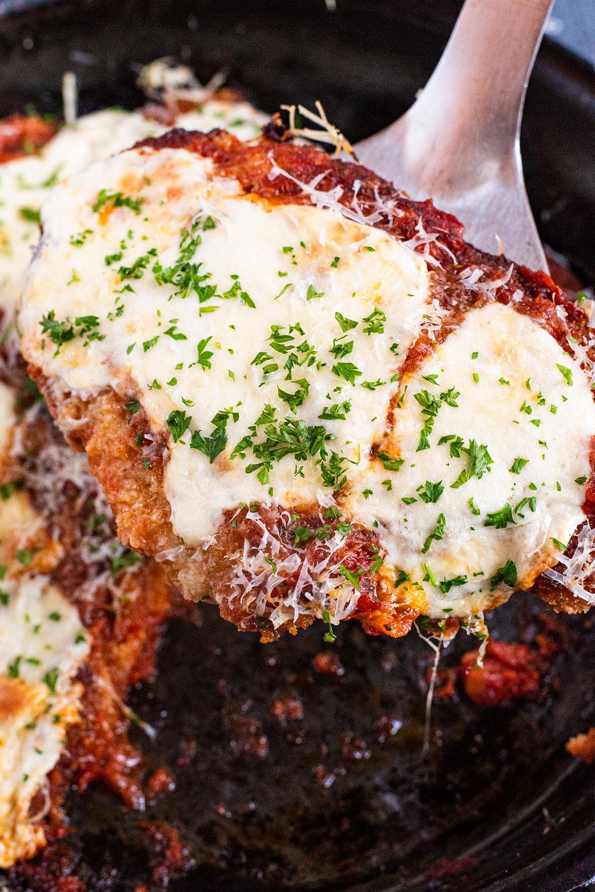 A spatula holding a piece of cheesy chicken parmesan out of a cast iron skillet.