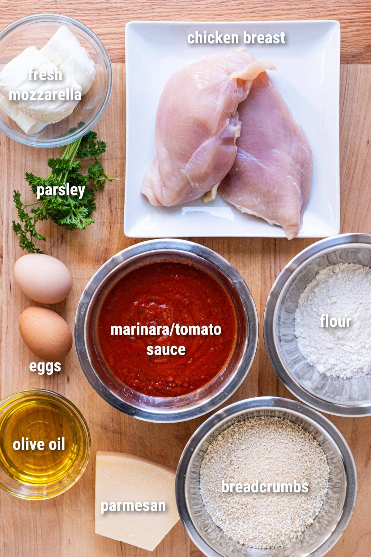 Raw, uncooked chicken parmesan ingredients laid out on a wooden cutting board.
