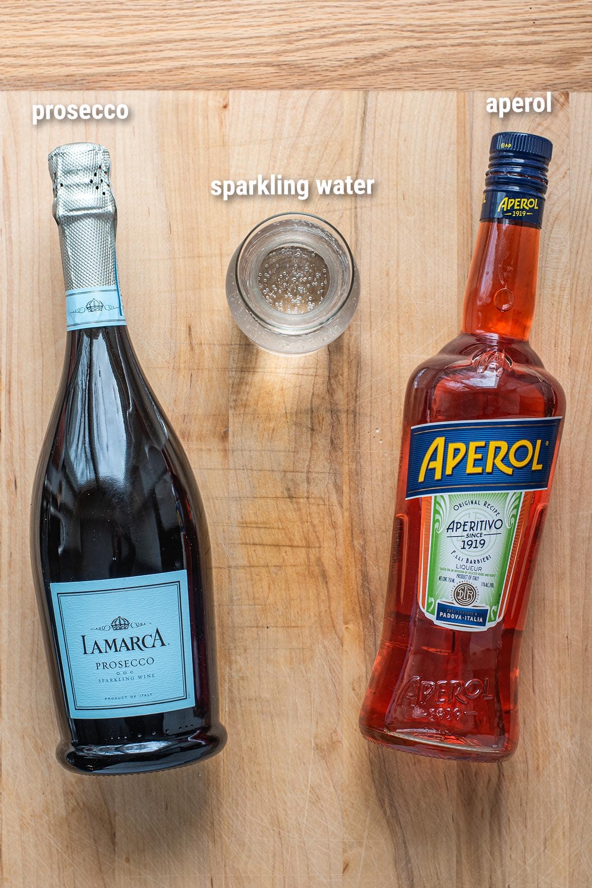 Aperol Spritz cocktail ingredients laid out on a cutting board. Aperol, Prosecco, and Sparkling water.
