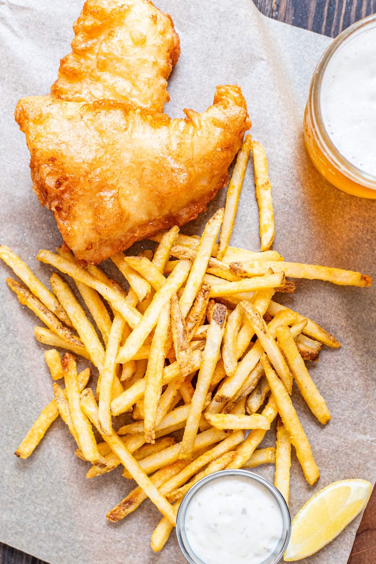 Fried beer-battered cod on a wooden table served with French fries, lemon, and tartar sauce.