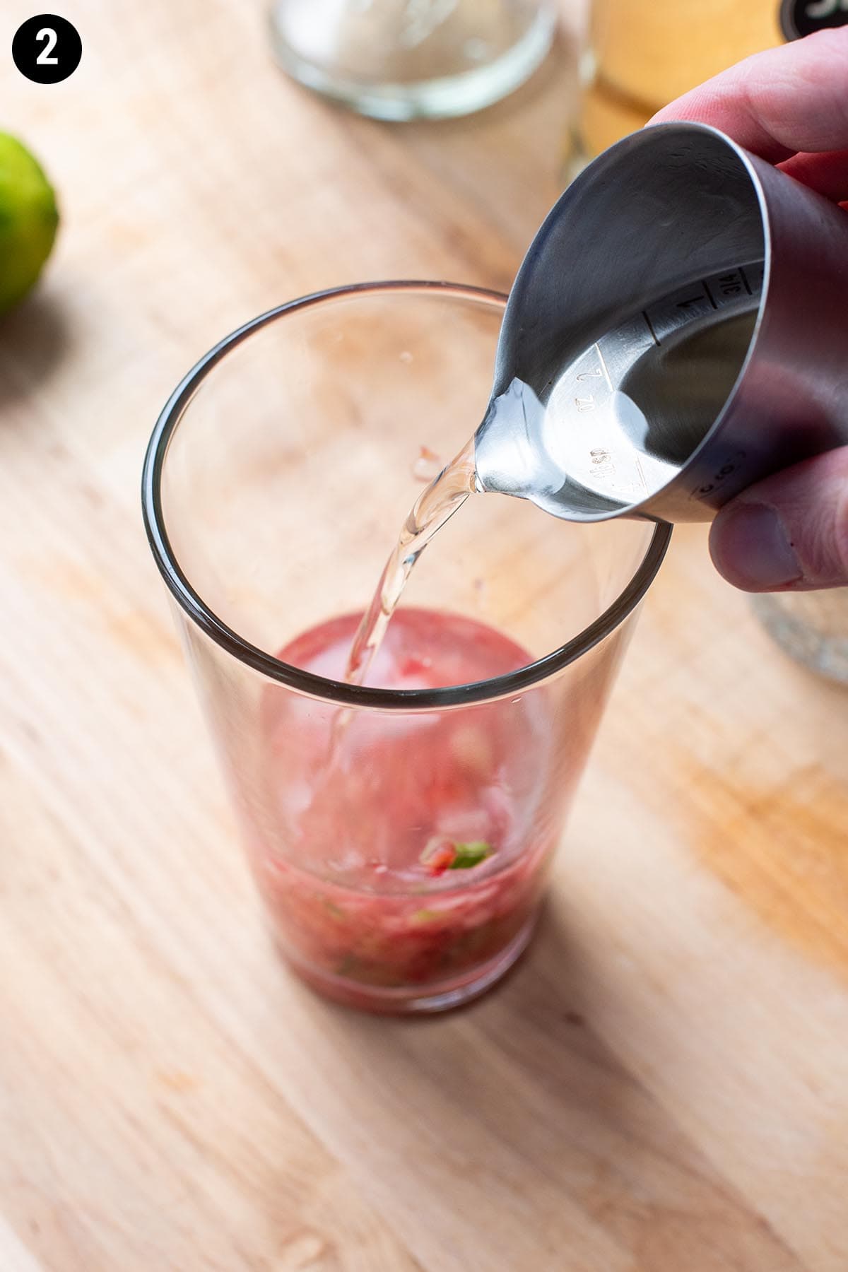 Pouring elderflower liqueur into a Boston shaker with muddled cucumber and strawberry.