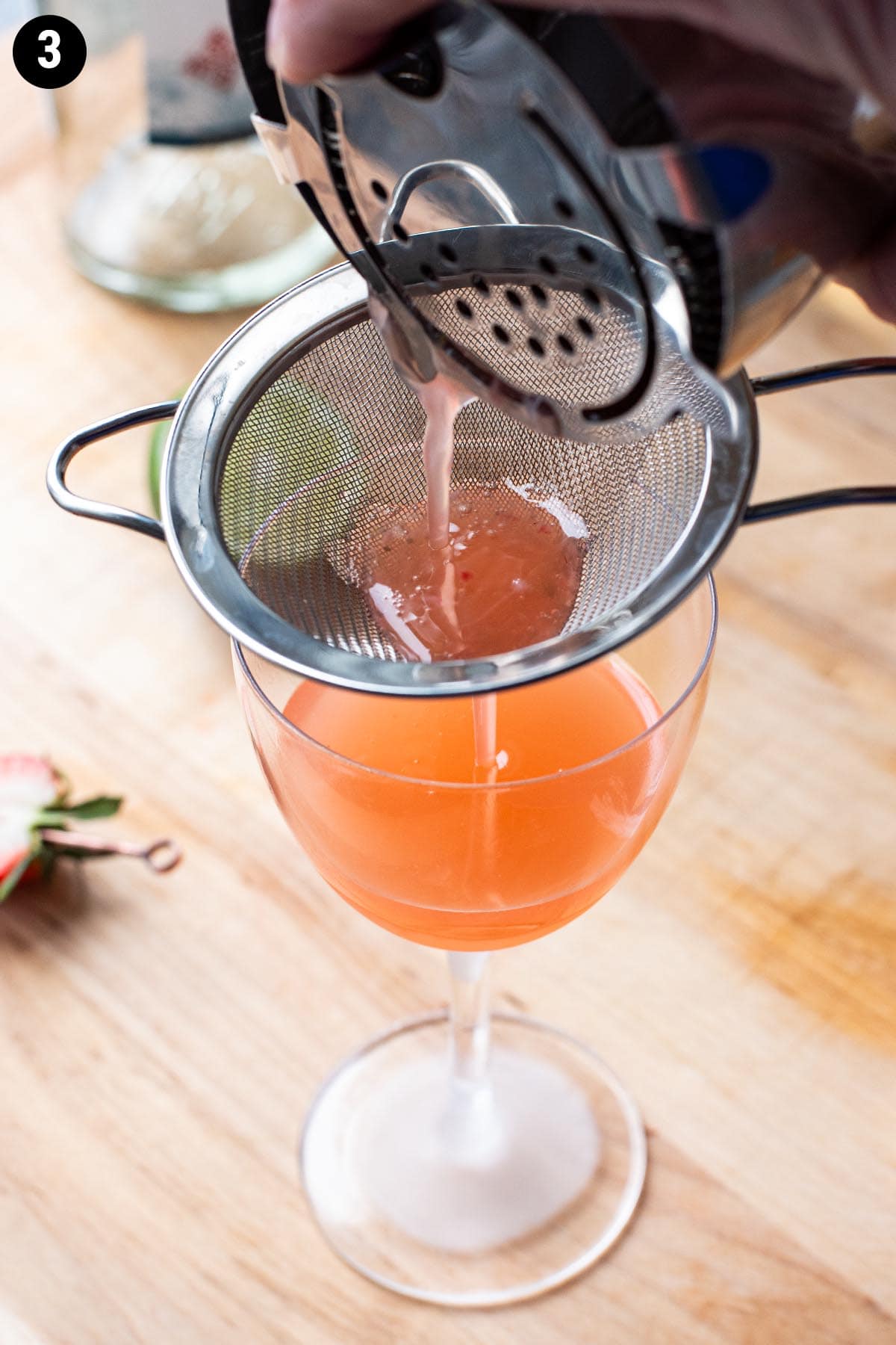Double straining a red hued cocktail into a chilled Nick and Nora glass through a fine mesh strainer.