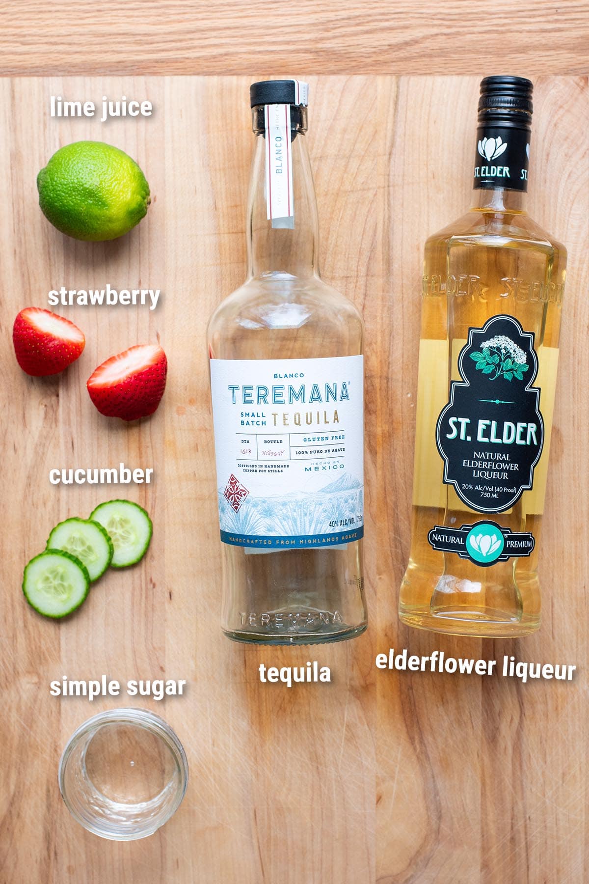 Ingredients to make a strawberry infused tequila martini laid out on a wooden cutting board.