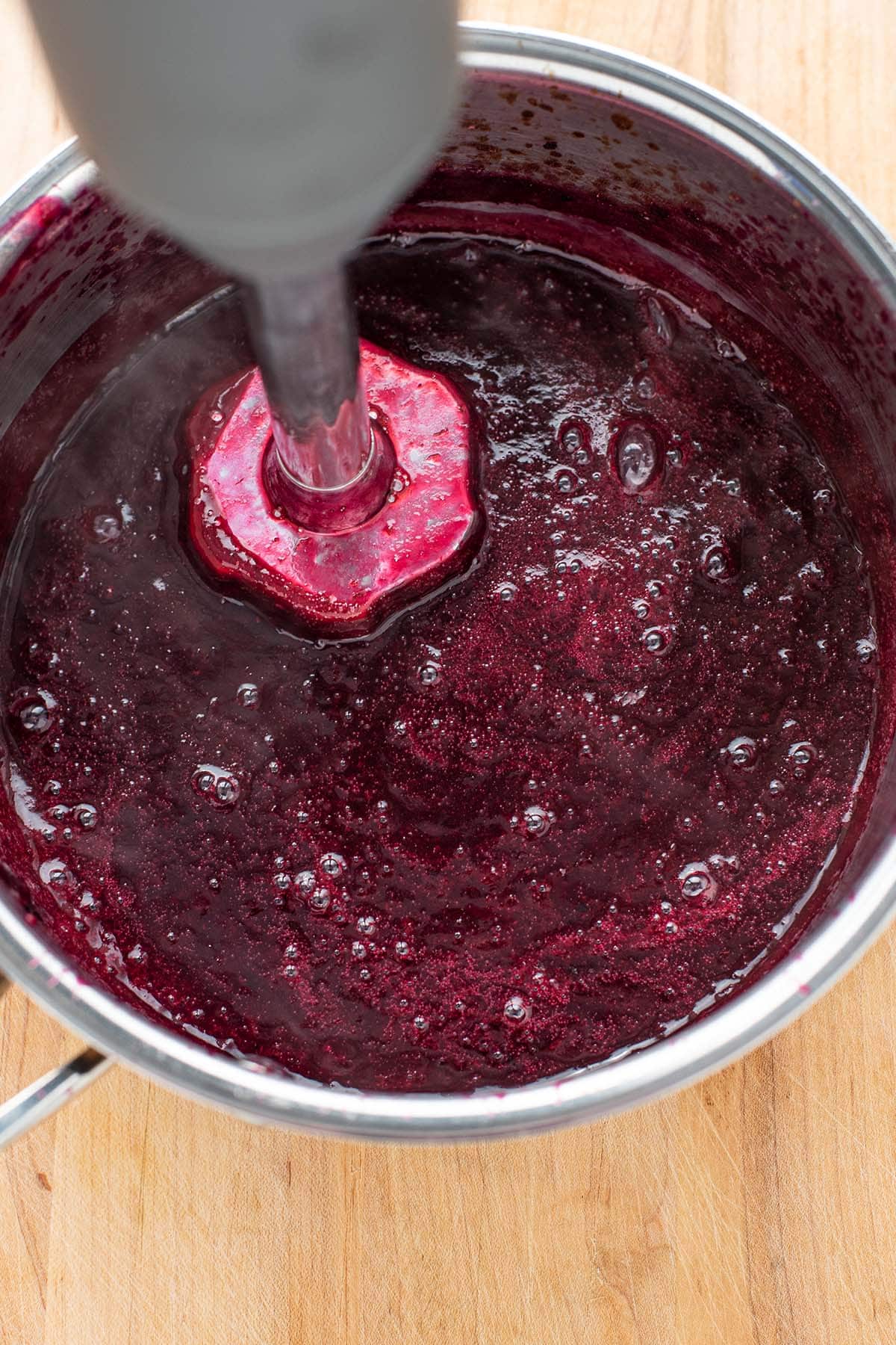 An immersion blender blending blueberries, red wine, sugar, and salt in a stainless saucepan.