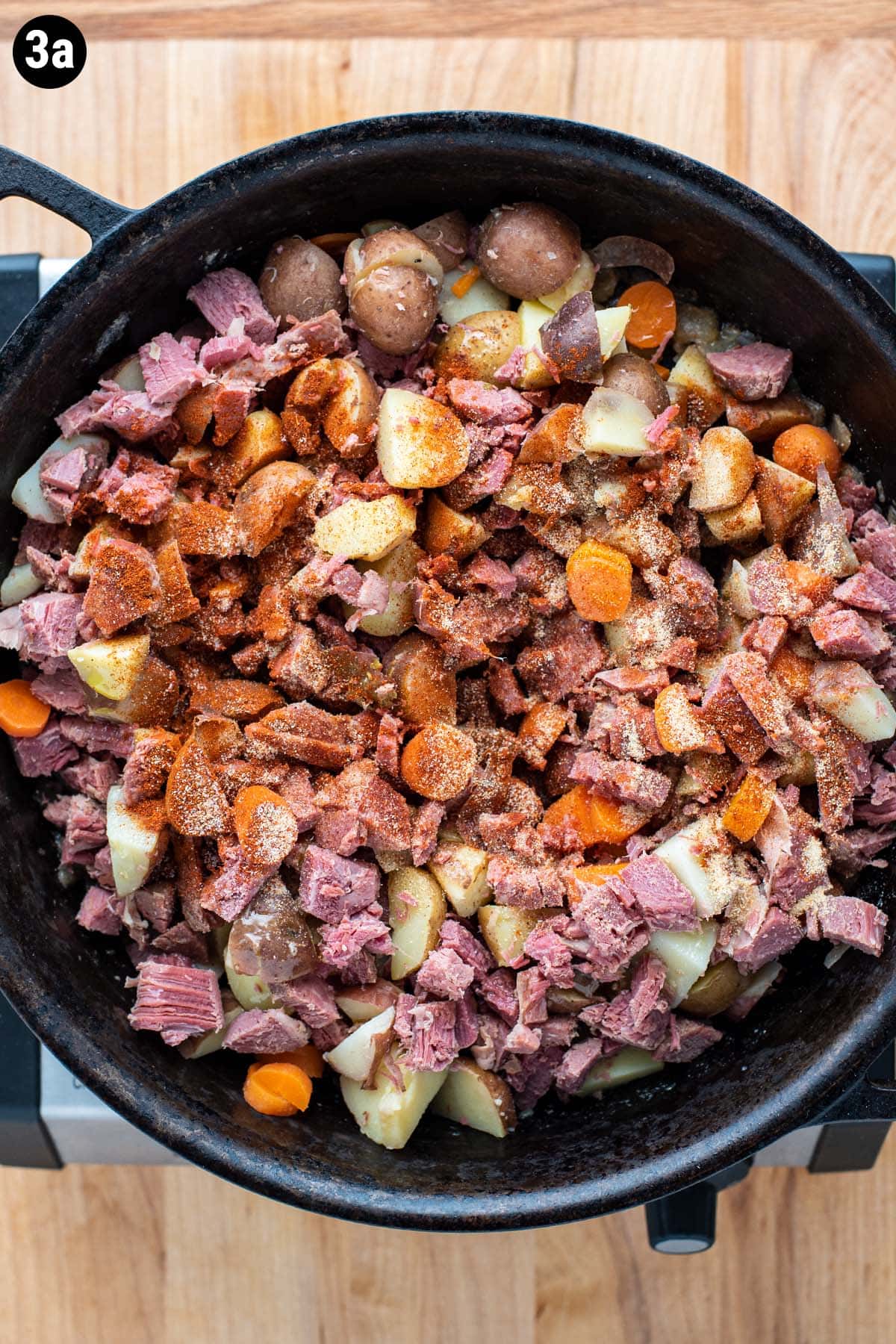Chopped carrots, potatoes, onion, and corned beef in a cast iron skillet topped with spices.
