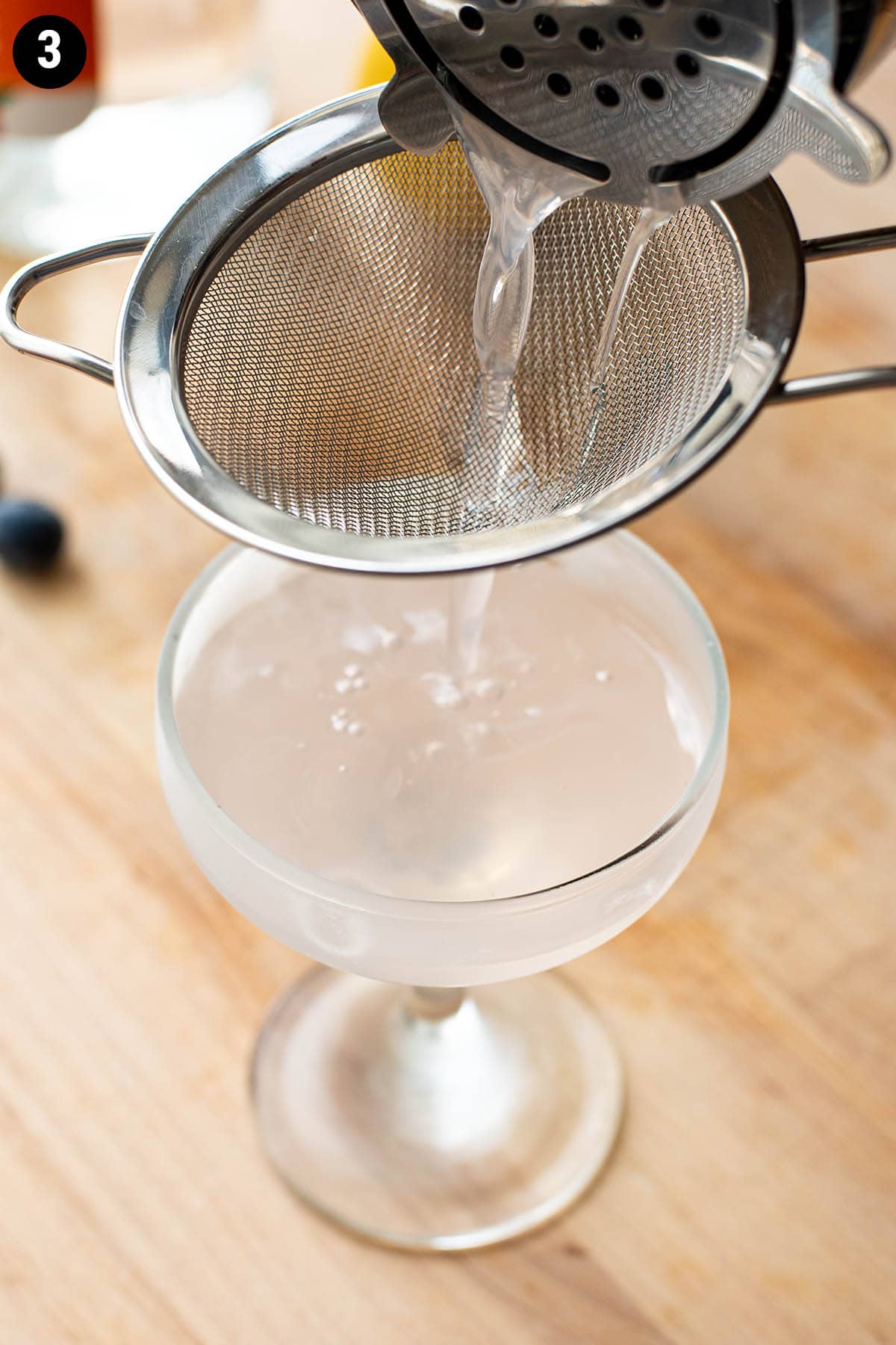 Double straining a blueberry martini into a chilled coupe glass.
