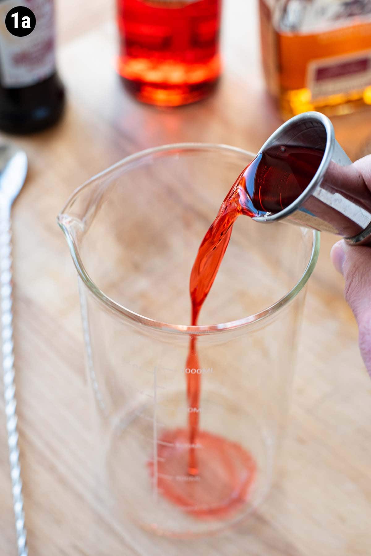 Pouring Campari into a glass mixing container with a double jigger.
