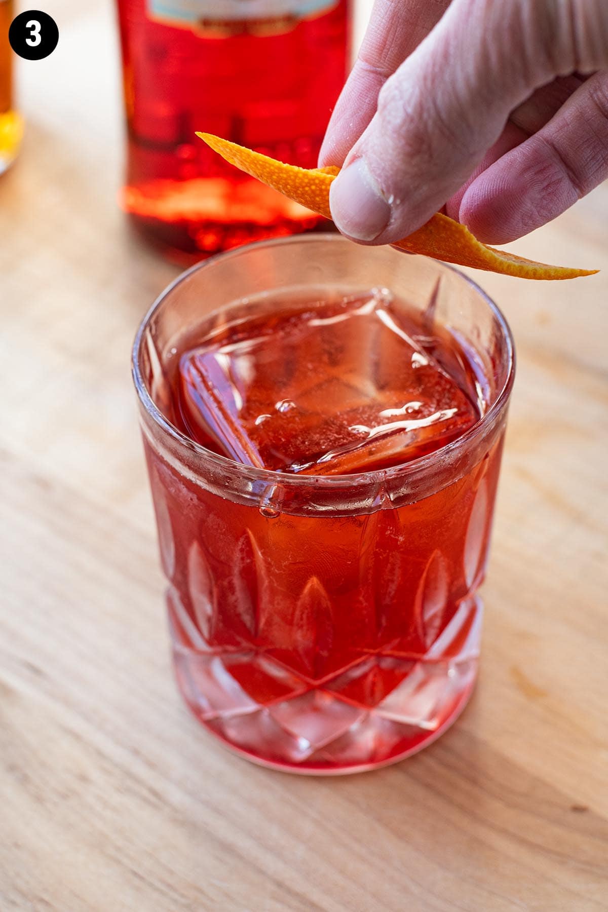 A hand holding an orange peel over a Boulevardier cocktail, expressing the oils over the drink.