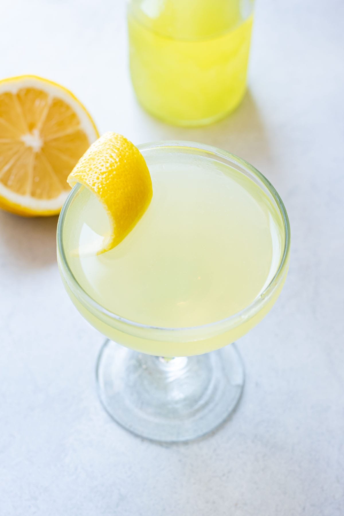 A pale yellow limoncello martini on a marble table garnished with a fancy lemon peel.
