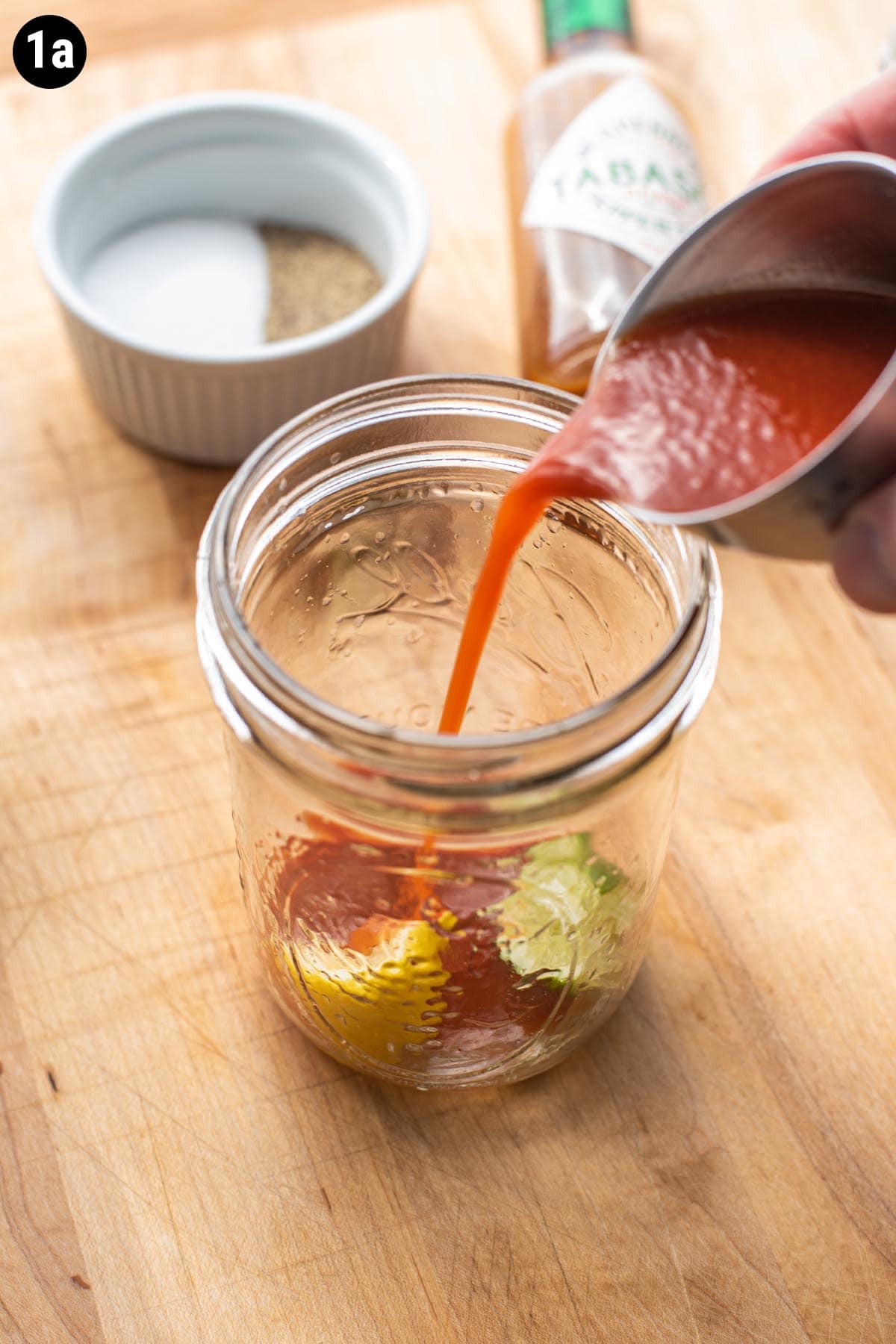 A hand pouring a double jigger of tomato juice into a glass with a lemon and lime wedge.