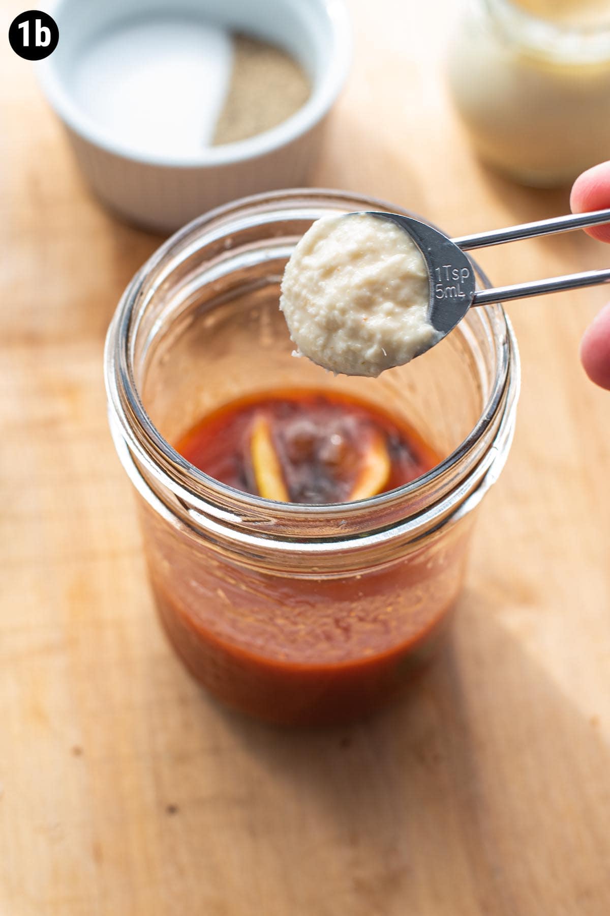A teaspoon of prepared horseradish being spooned into a mason jar of tomato juice.