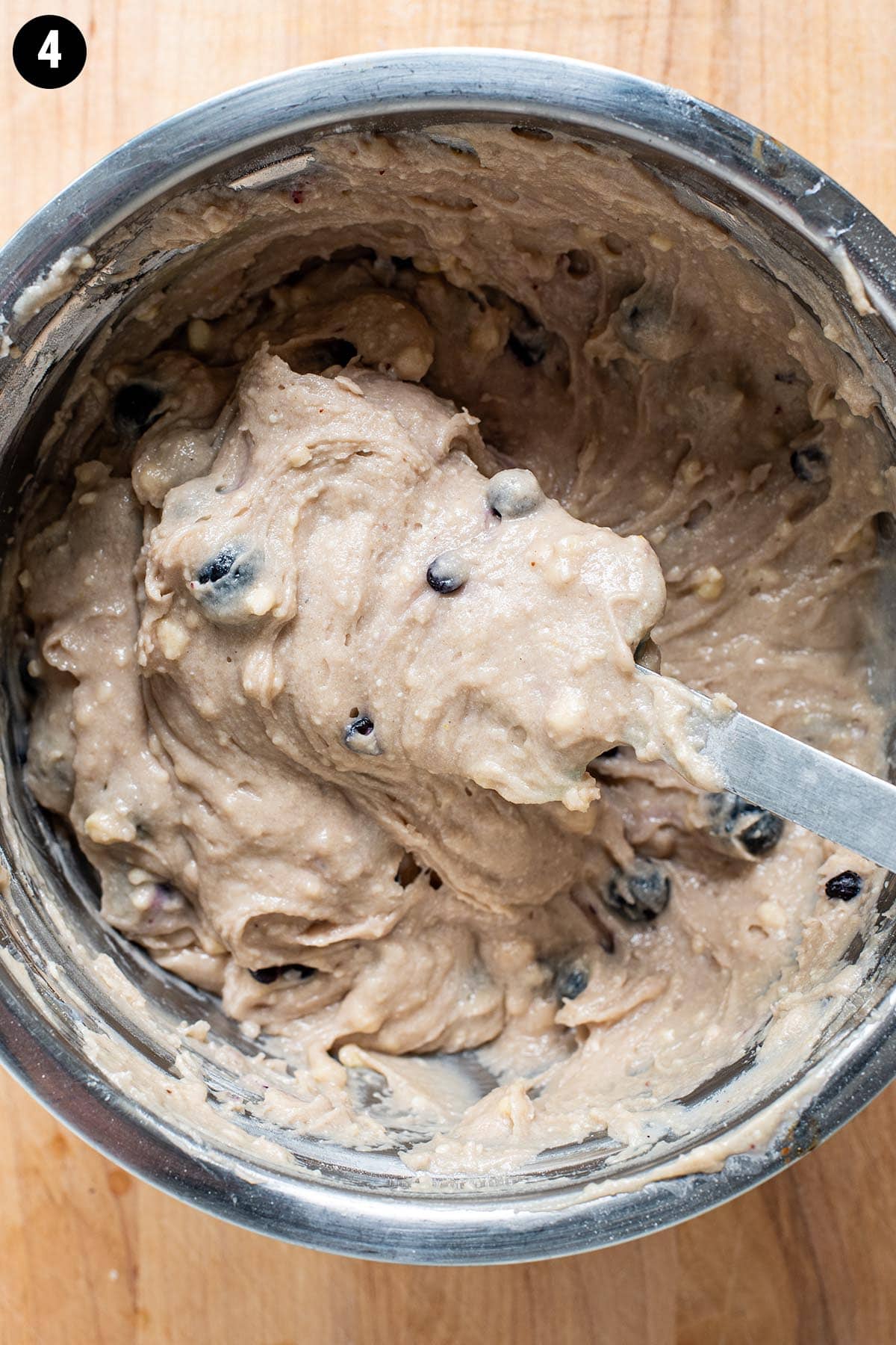 A rubber spatula folding blueberries into a thick blueberry muffin batter in a stainless steel bowl.
