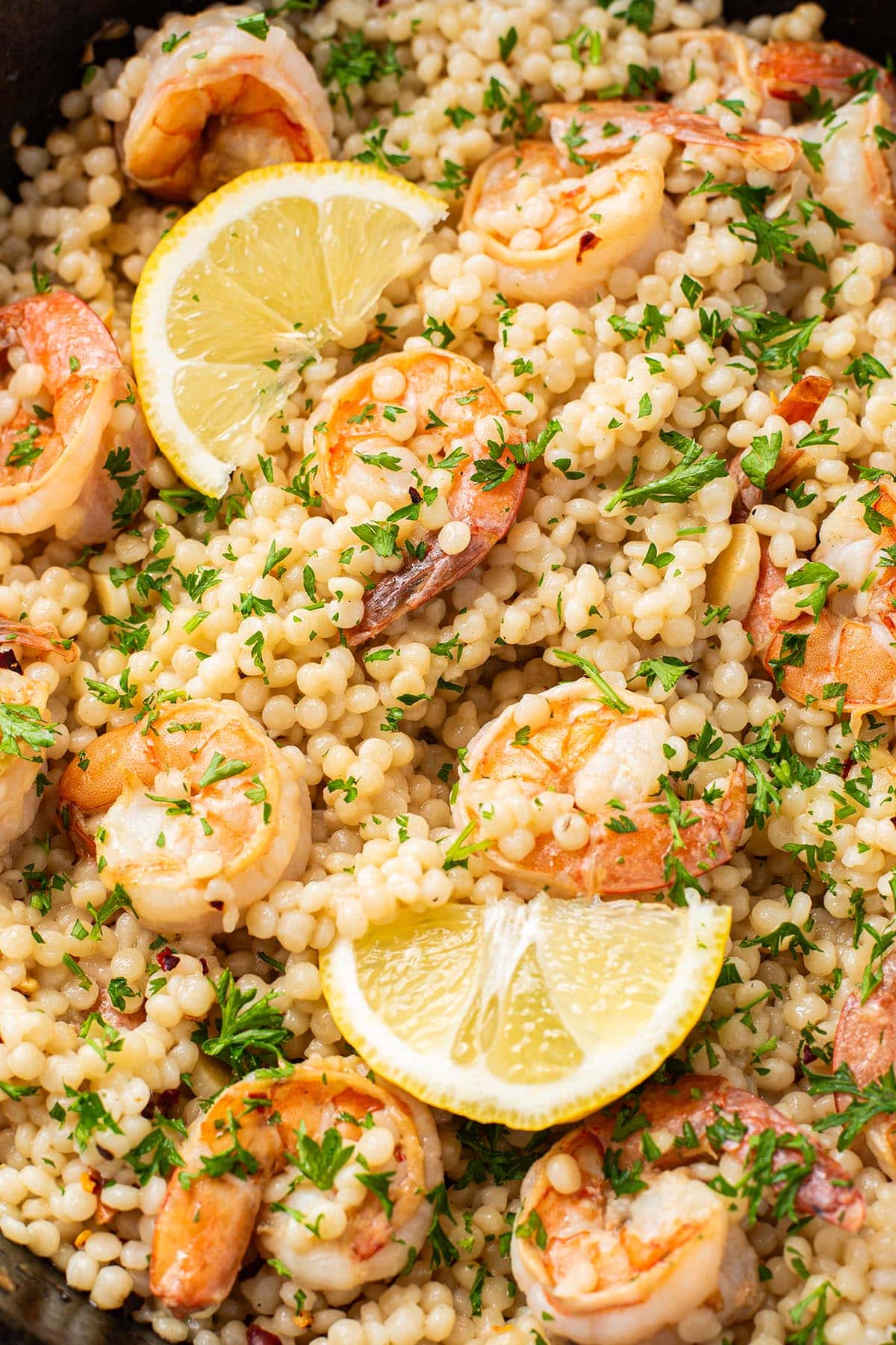Shrimp scampi in a skillet made with pearl couscous and topped with lemon wedges and fresh parsley.