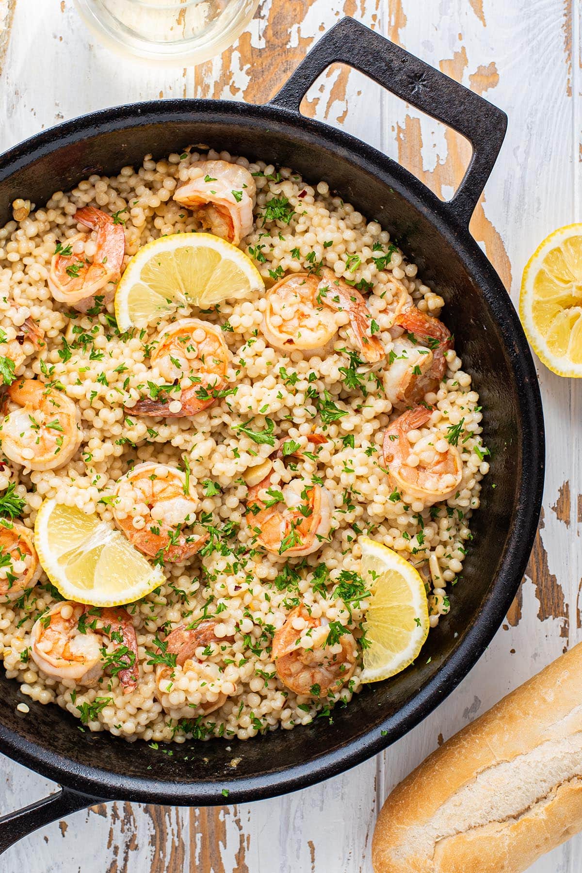Shrimp scampi with pearl couscous in a skillet on a wooden white table with bread and white wine.