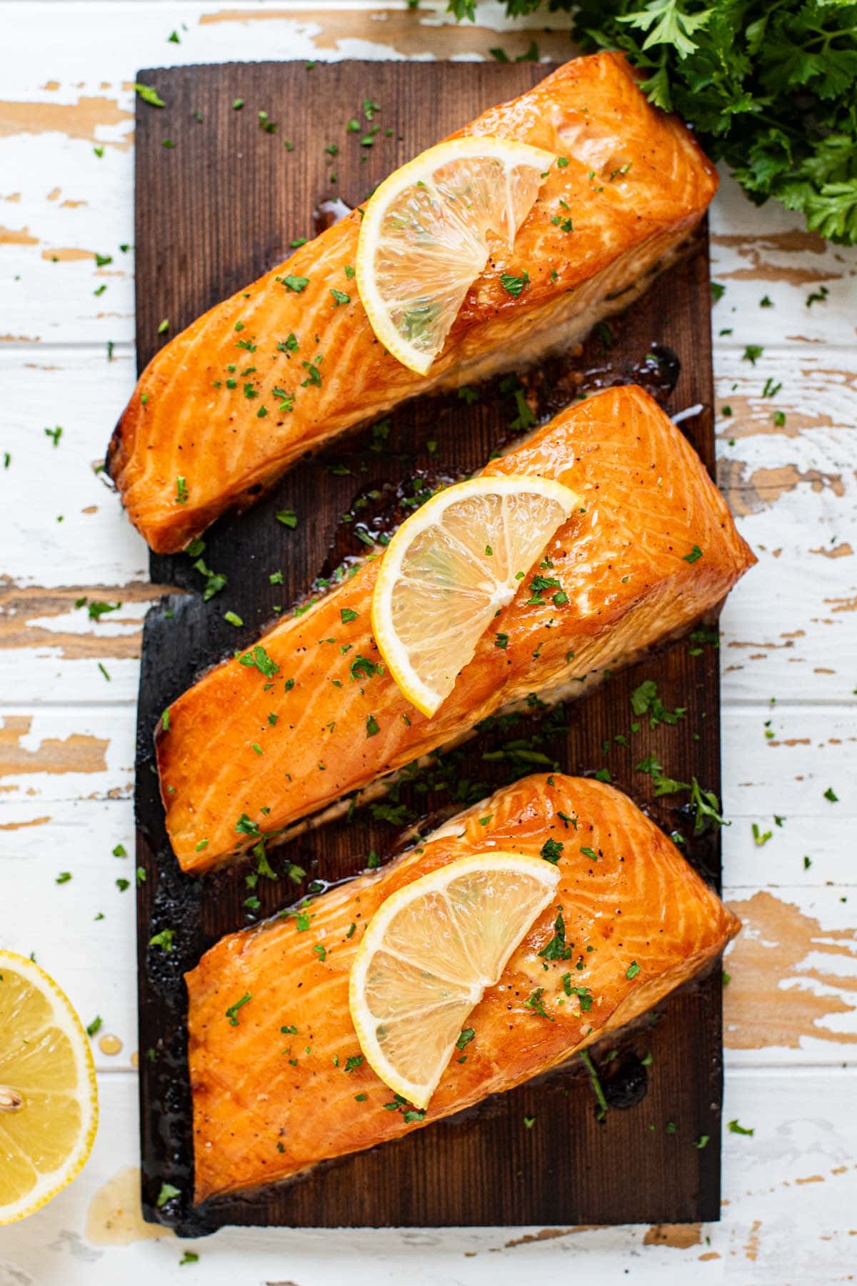Three golden salmon filets on a cedar plank topped with fresh parsley and lemon wedges.