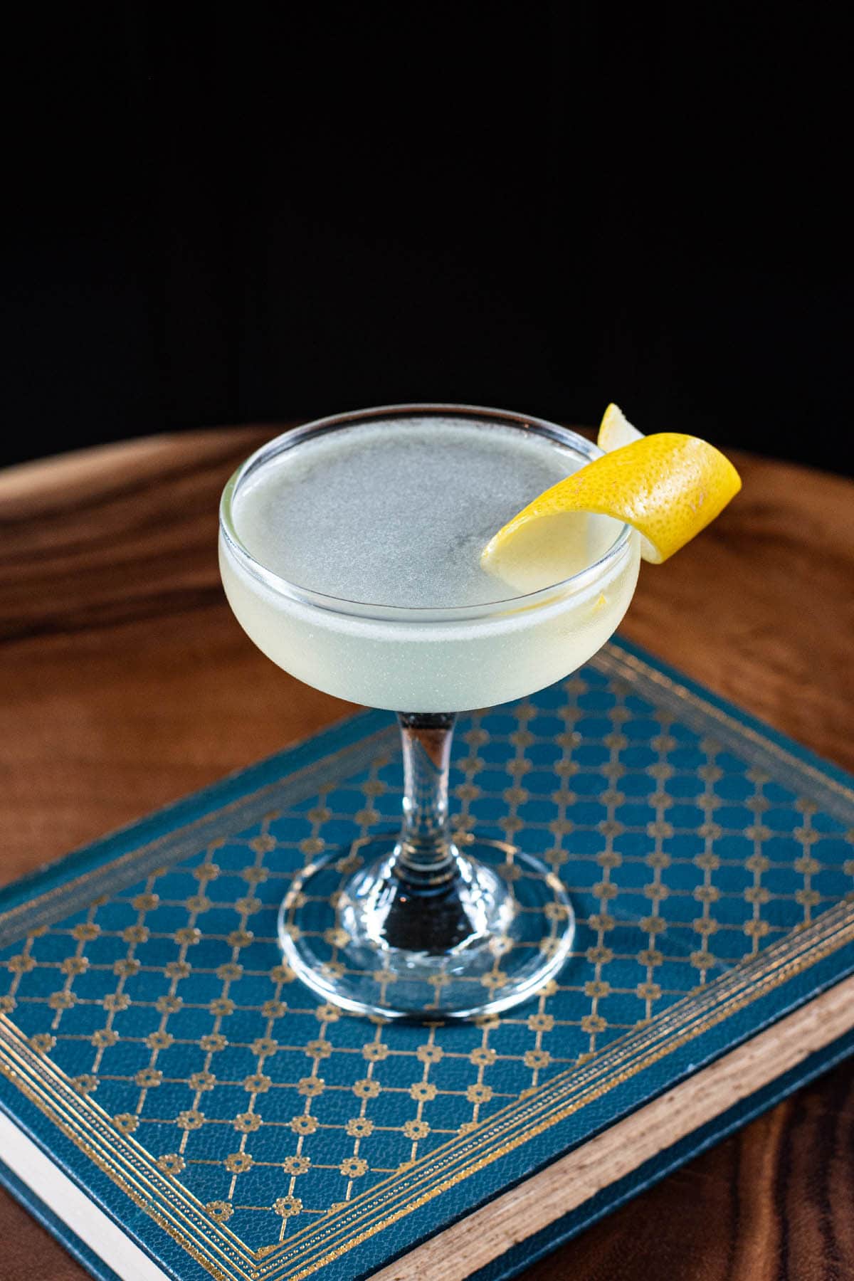 A pale yellow Corpse Reviver No. 2 cocktail garnished with a lemon twist, sitting on top of a blue book.