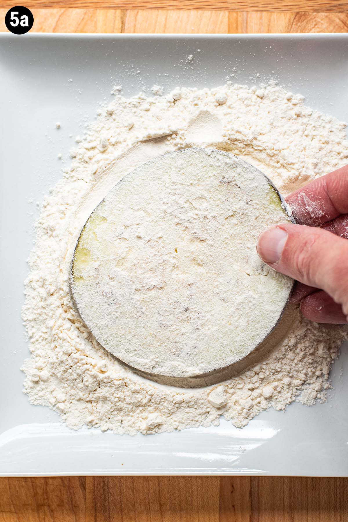 A hand dredging a slice of uncooked eggplant in all-purpose flour.