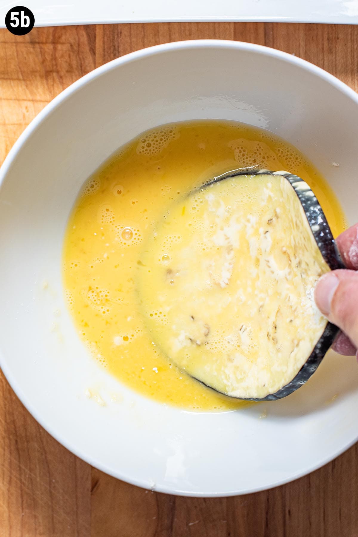 A hand dredging a slice of uncooked eggplant in whisked egg.