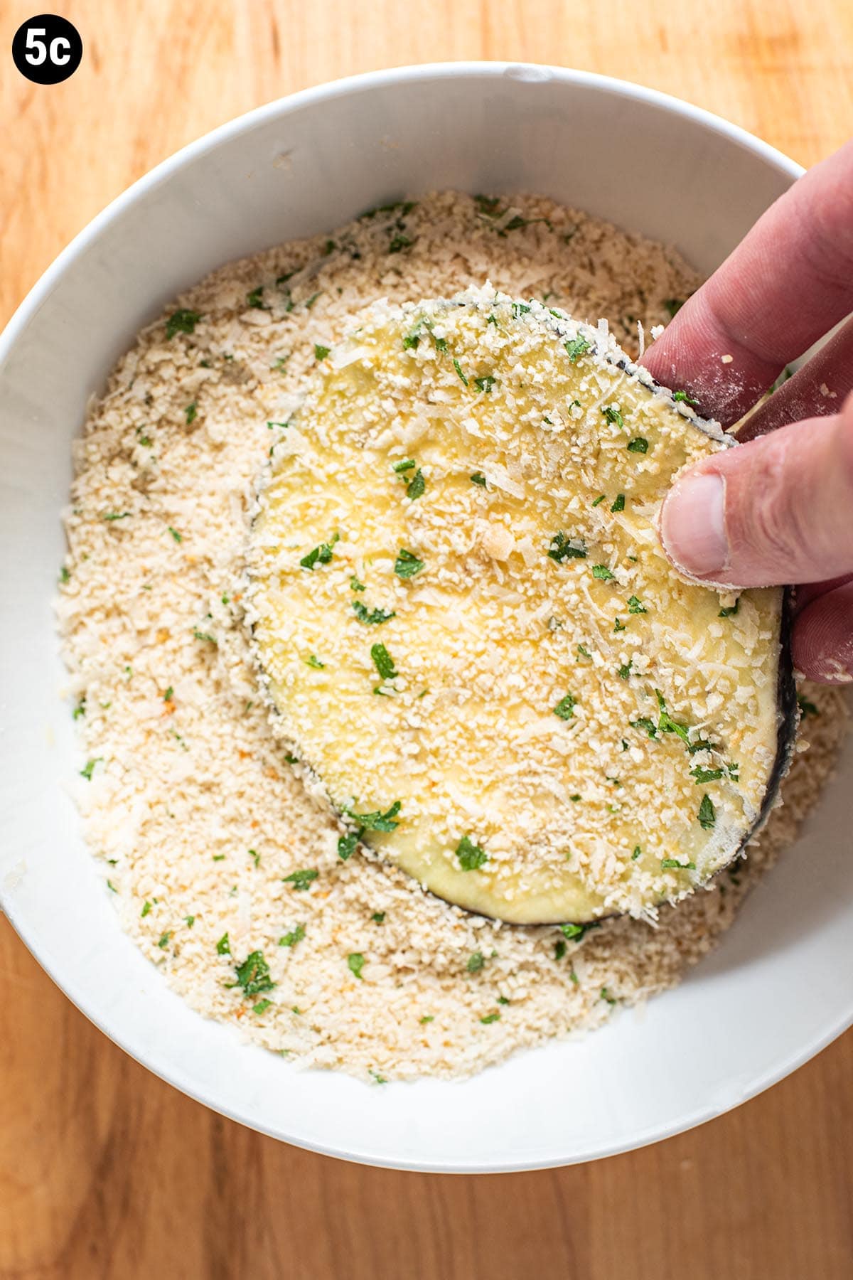 A hand dredging a slice of uncooked eggplant in a mix of breadcrumbs and parmesan cheese.