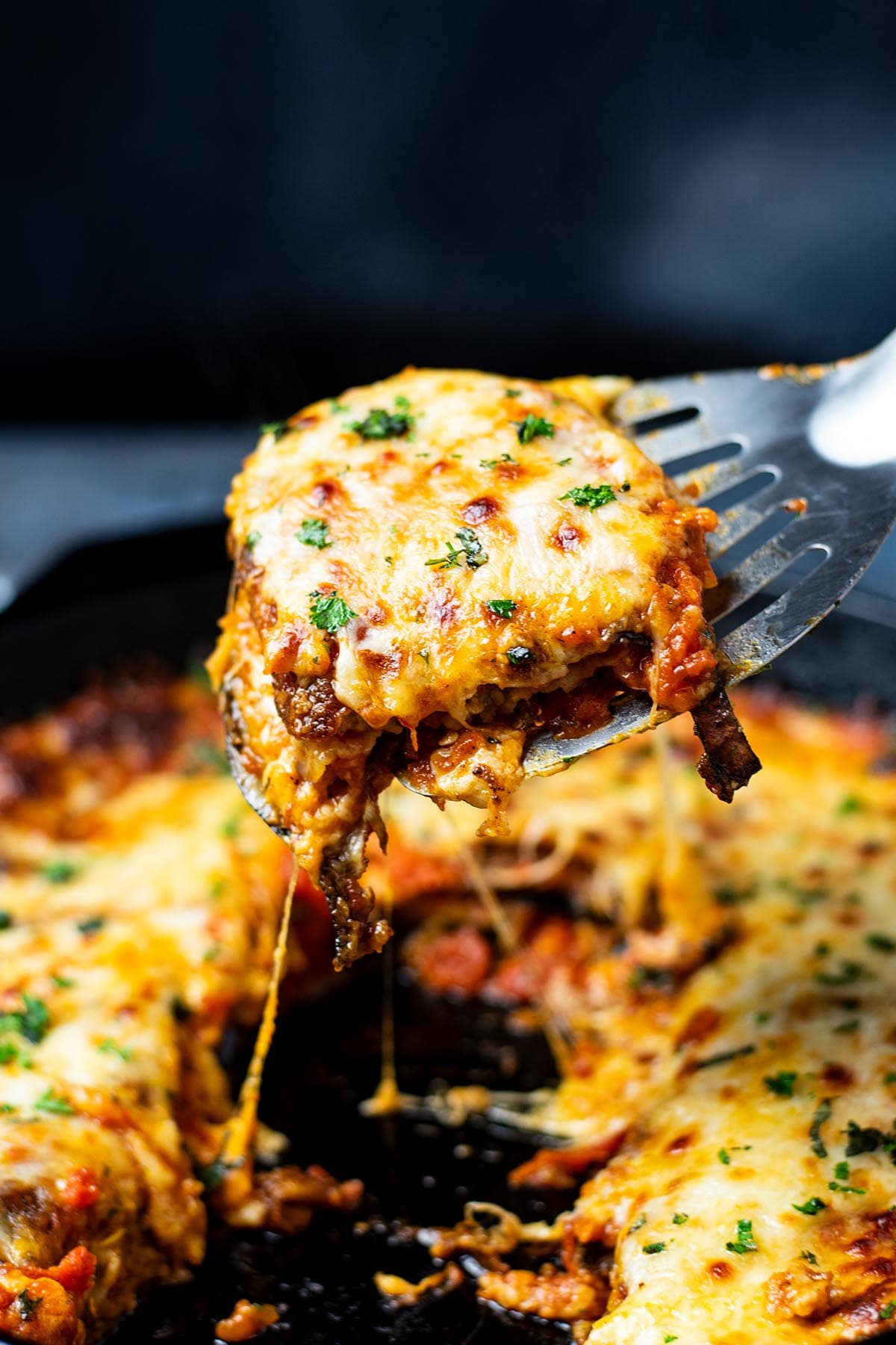 A metal spatula removing a slice of crispy golden eggplant parmesan from a cast iron skillet.