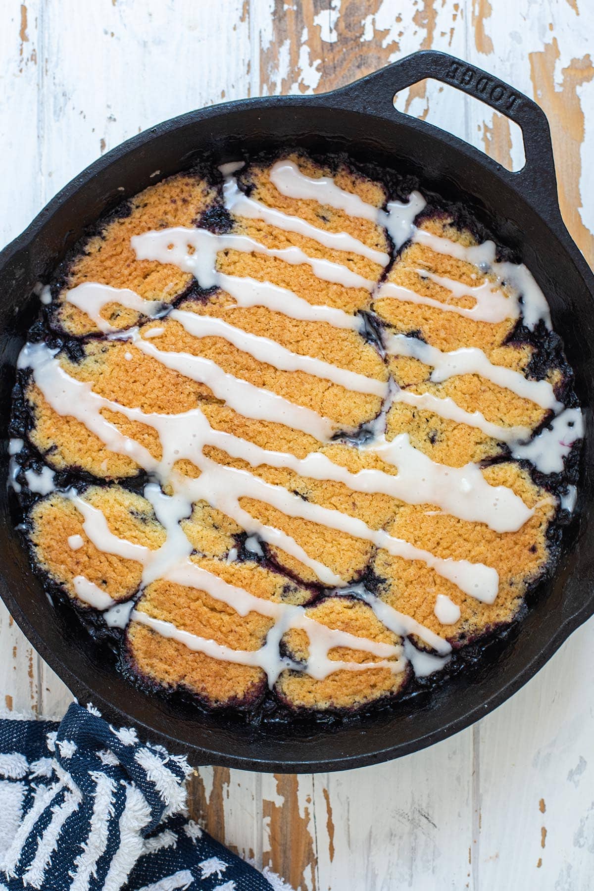 Golden baked blueberry cobbler in a cast iron skillet topped with an almond frosting glaze.