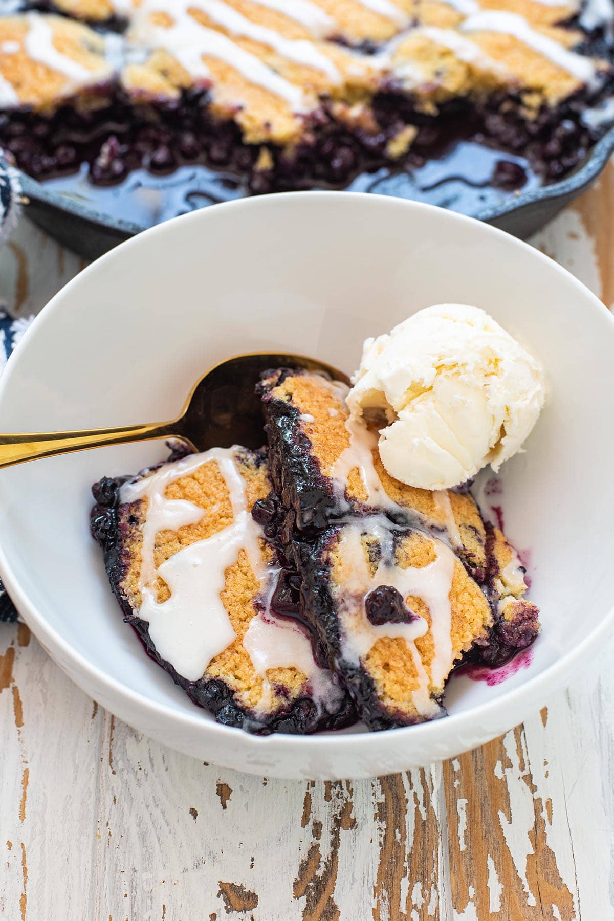 A bowl of golden blueberry cobbler topped with an almond frosting glaze and vanilla ice cream.