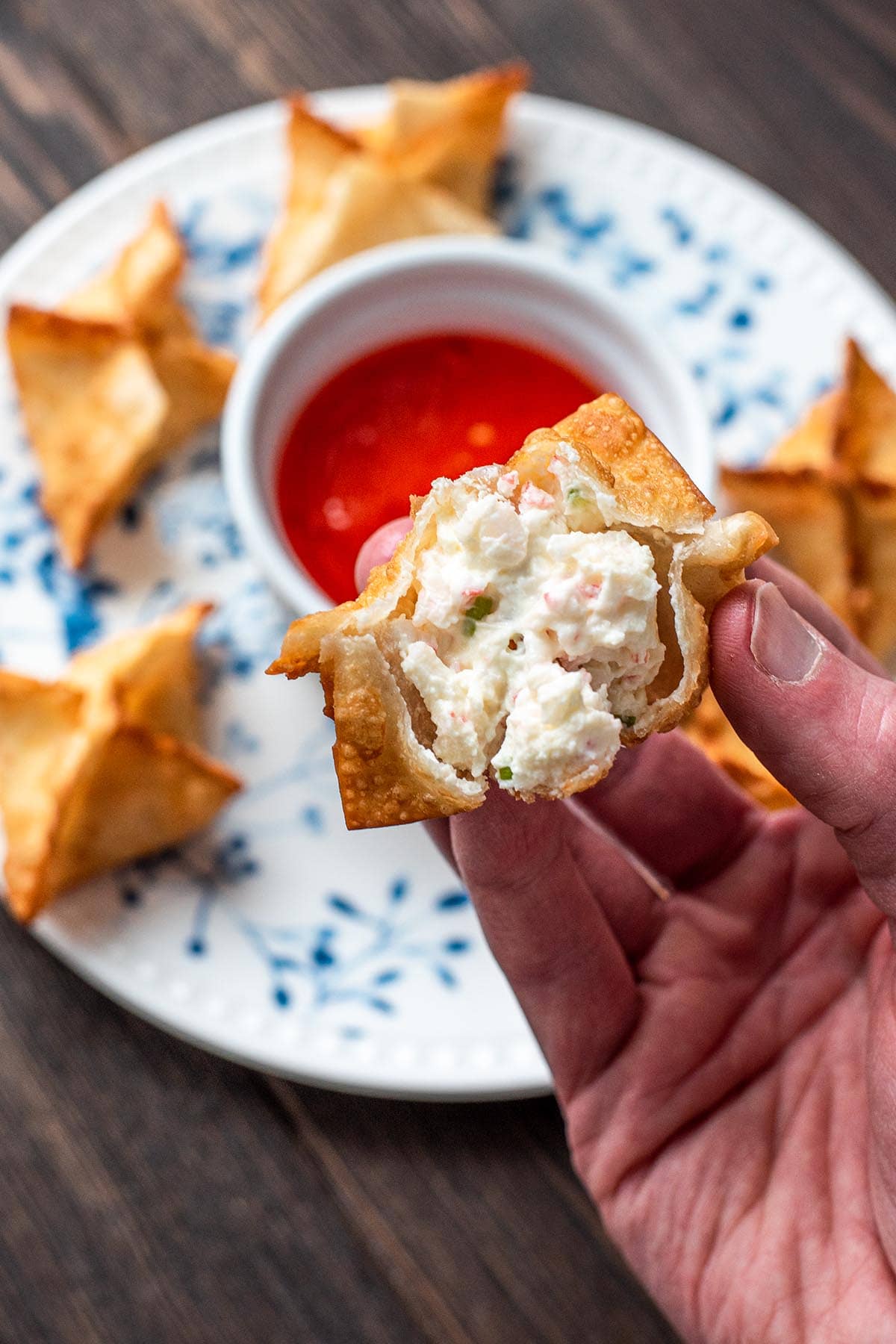 A hand holding a cooked crab rangoon, exposing the cream cheese filling.