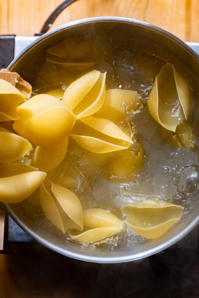 Uncooked jumbo pasta shells being dumped into a large pot of boiling water.