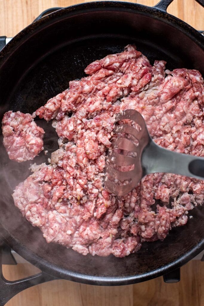 A metal spatula breaking up ground Italian sausage as it cooks in a cast iron skillet.