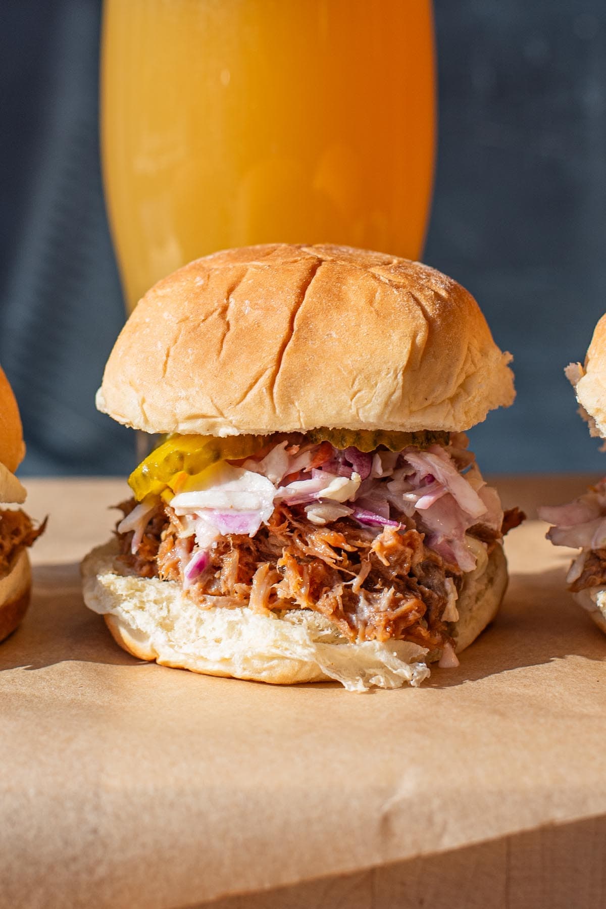 A BBQ pulled pork slider topped with coleslaw and pickles, pictured in front of a golden hazy beer.