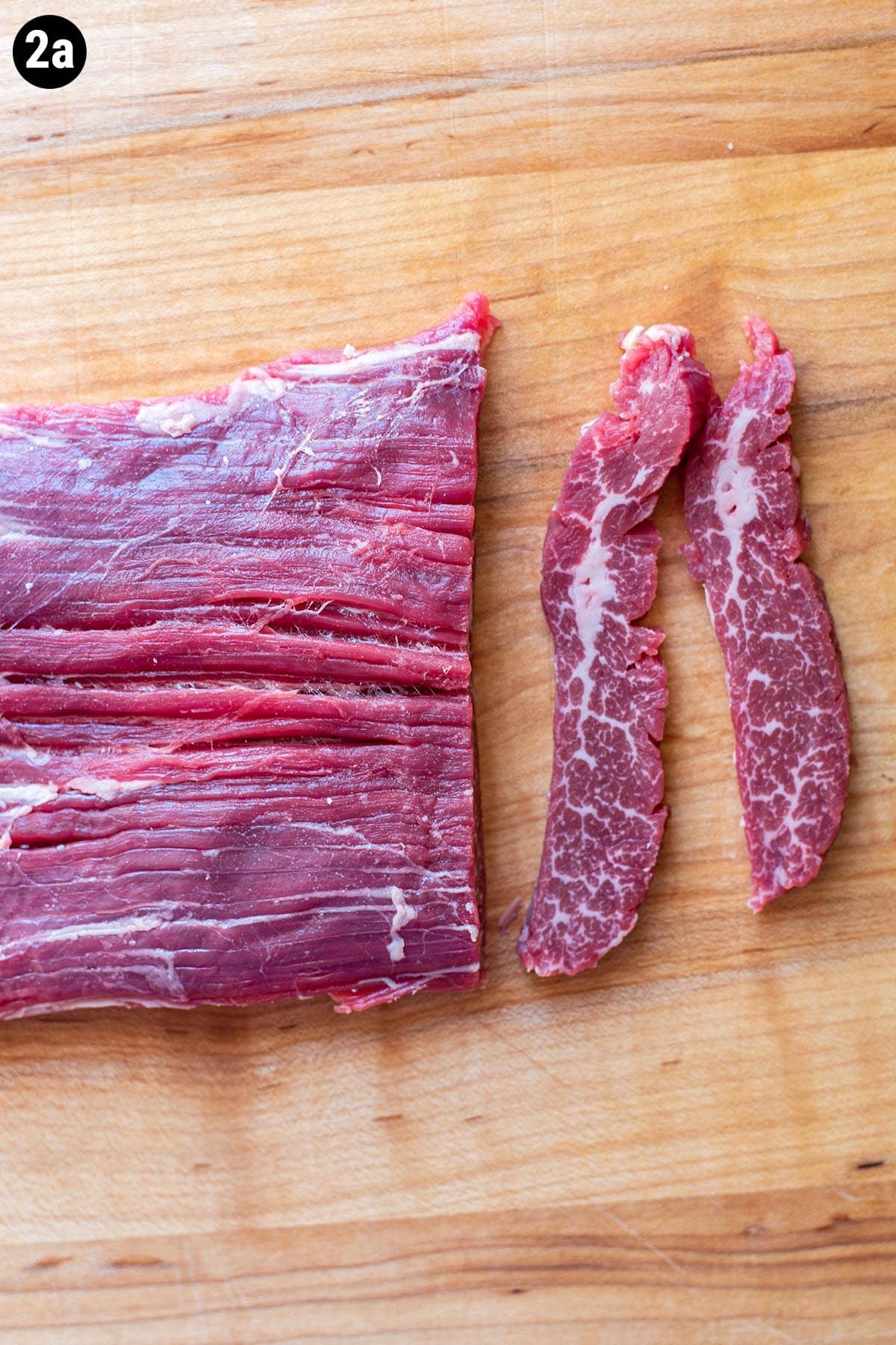 An uncooked piece of flank steak on a cutting board with two thin slices sliced off.