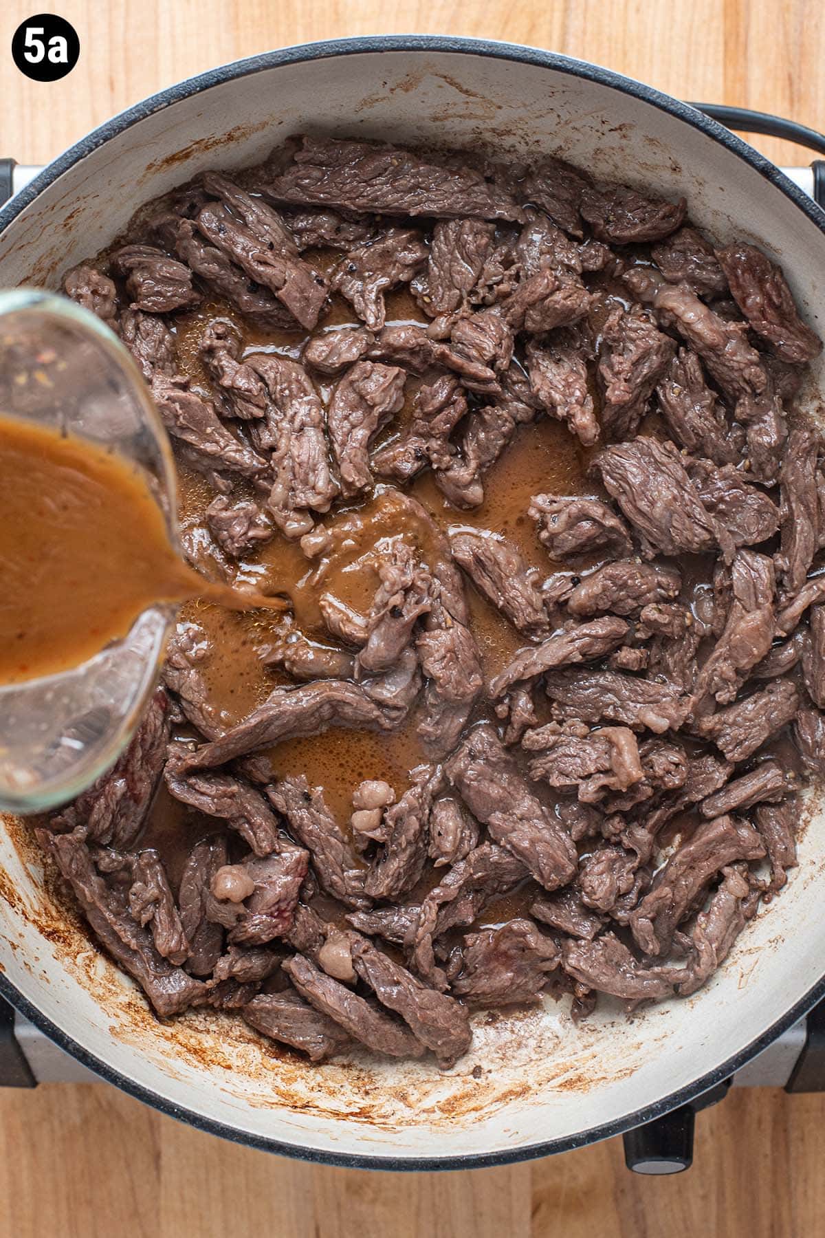 Cooked flank steak in a pan and a hand pouring in a beef and broccoli sauce before simmering.