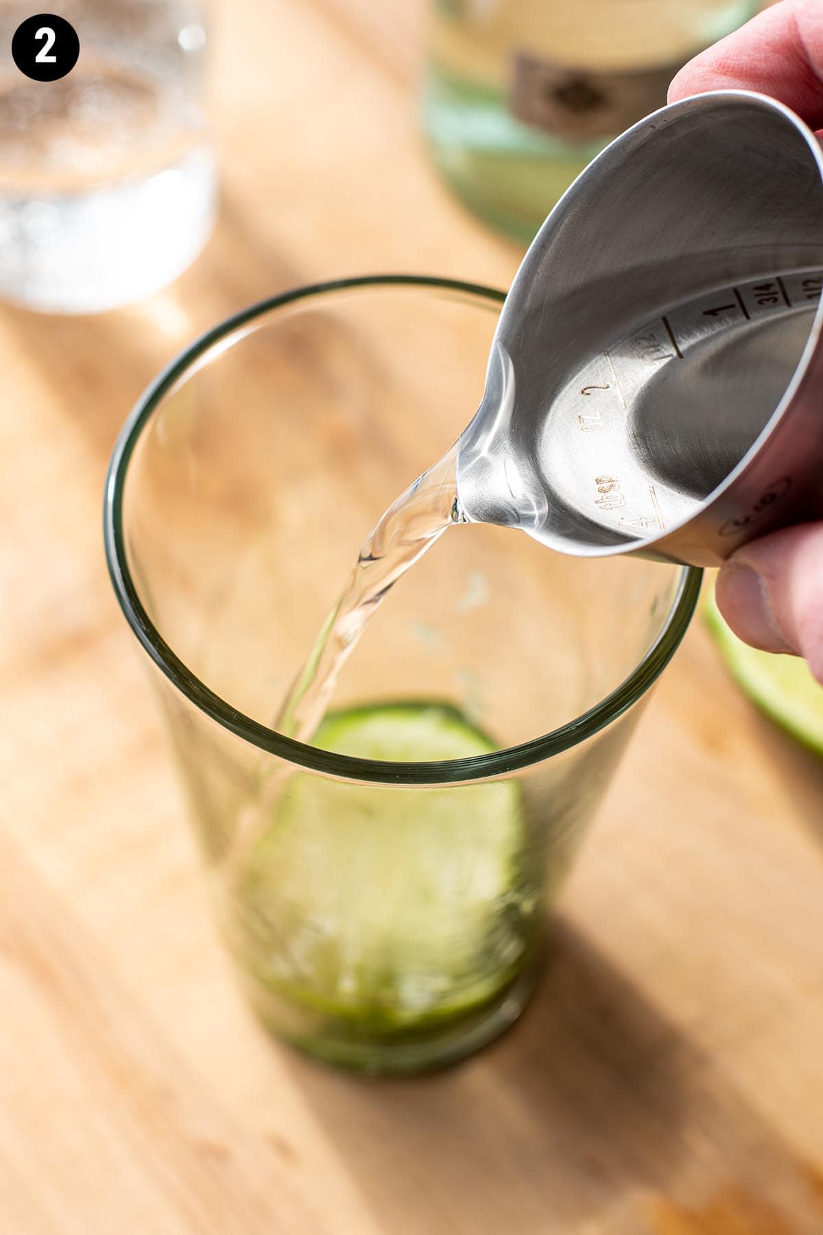 A jigger pouring silver rum into a mixing glass with muddled mint leaves and lime wedges.