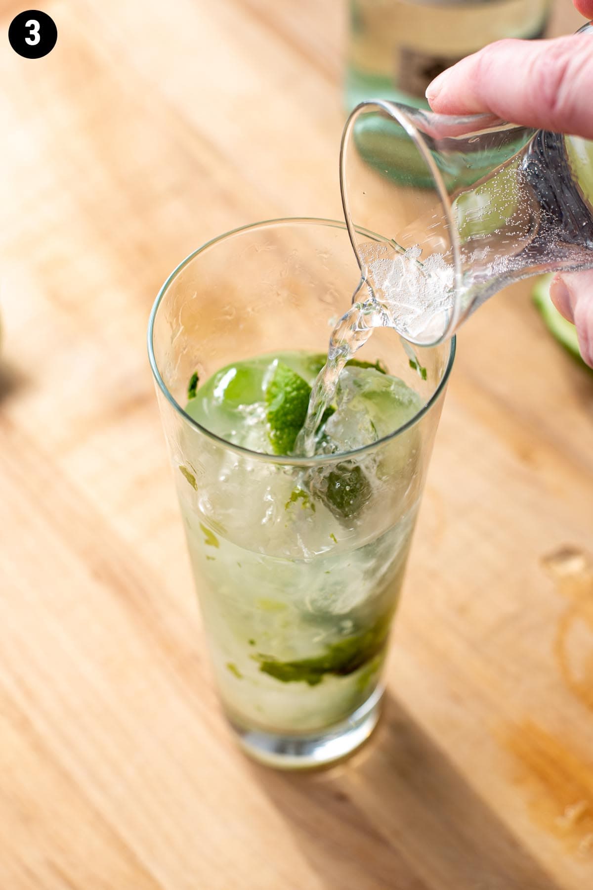 Pouring soda water into a tall glass holding a mojito cocktail garnished with mint leaves and lime wedges.