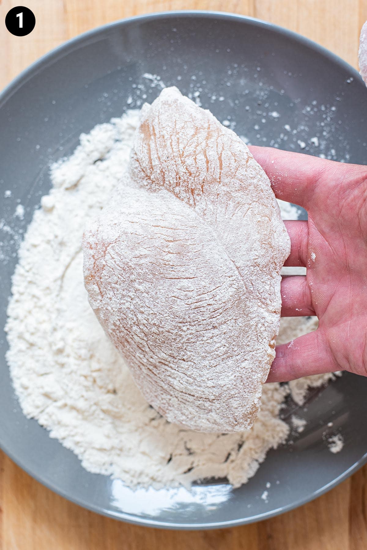 A hand holding a raw piece of chicken that has been dredged in all-purpose flour