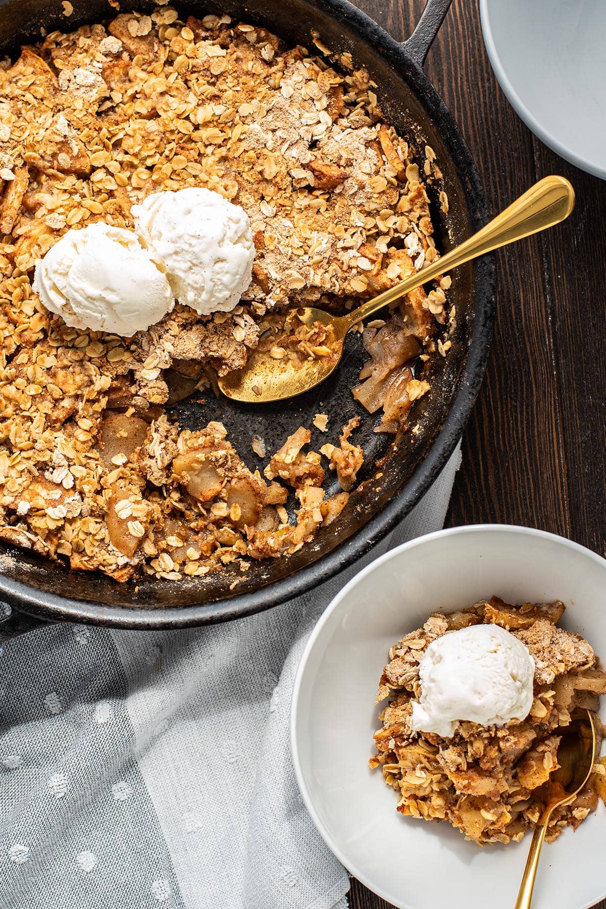 Golden apple crisp in a cast iron skillet topped with two scoops of vanilla ice cream and a serving removed.