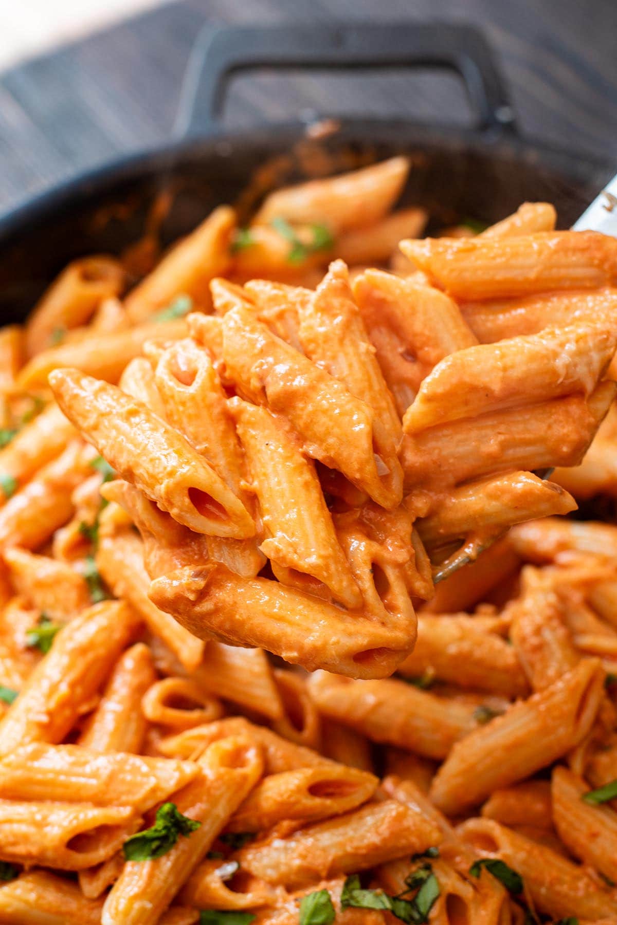 A metal spoon lifting a heaping spoonful of creamy penne alla vodka out of a cast iron skillet.