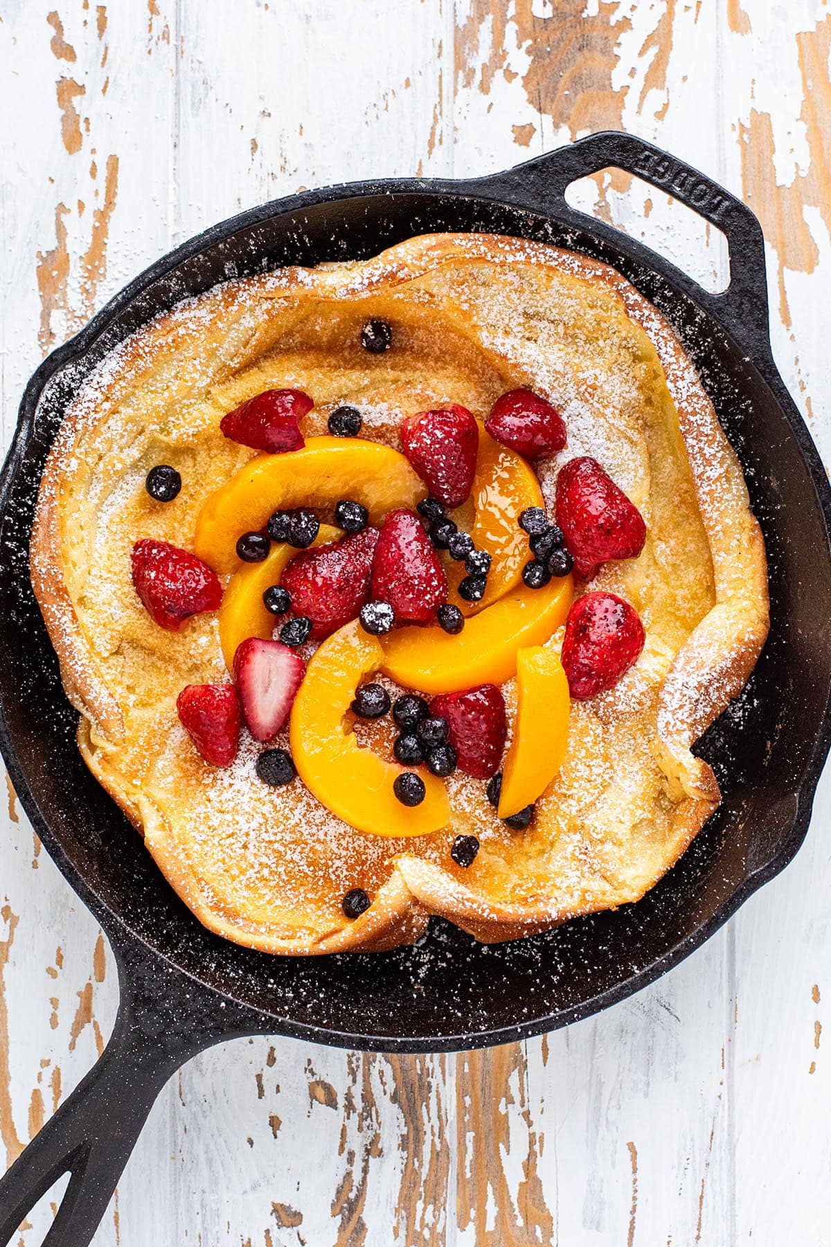 A golden Dutch baby in a cast iron skillet topped with strawberries, peaches, blueberries, maple syrup, and powdered sugar.