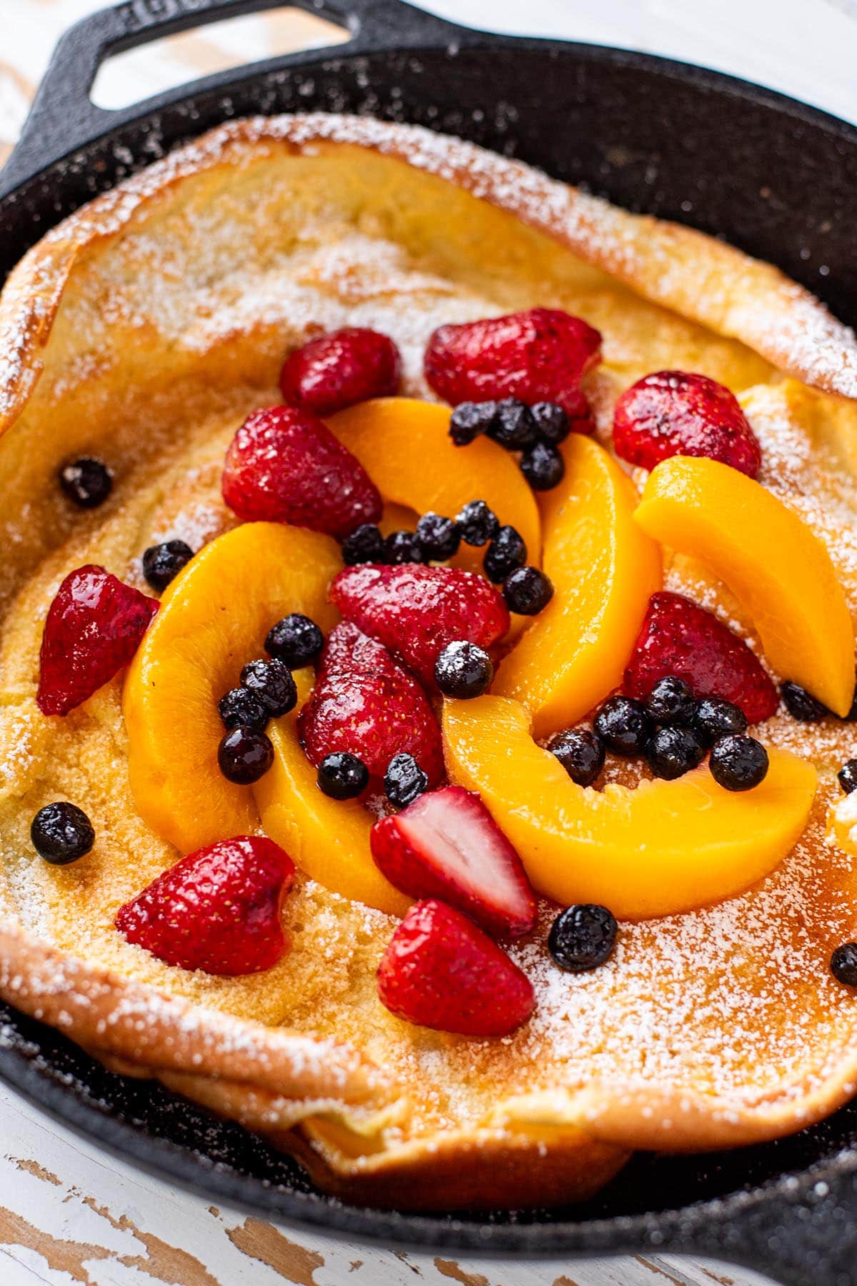 A golden Dutch baby in a cast iron skillet topped with strawberries, peaches, blueberries, maple syrup, and powdered sugar.