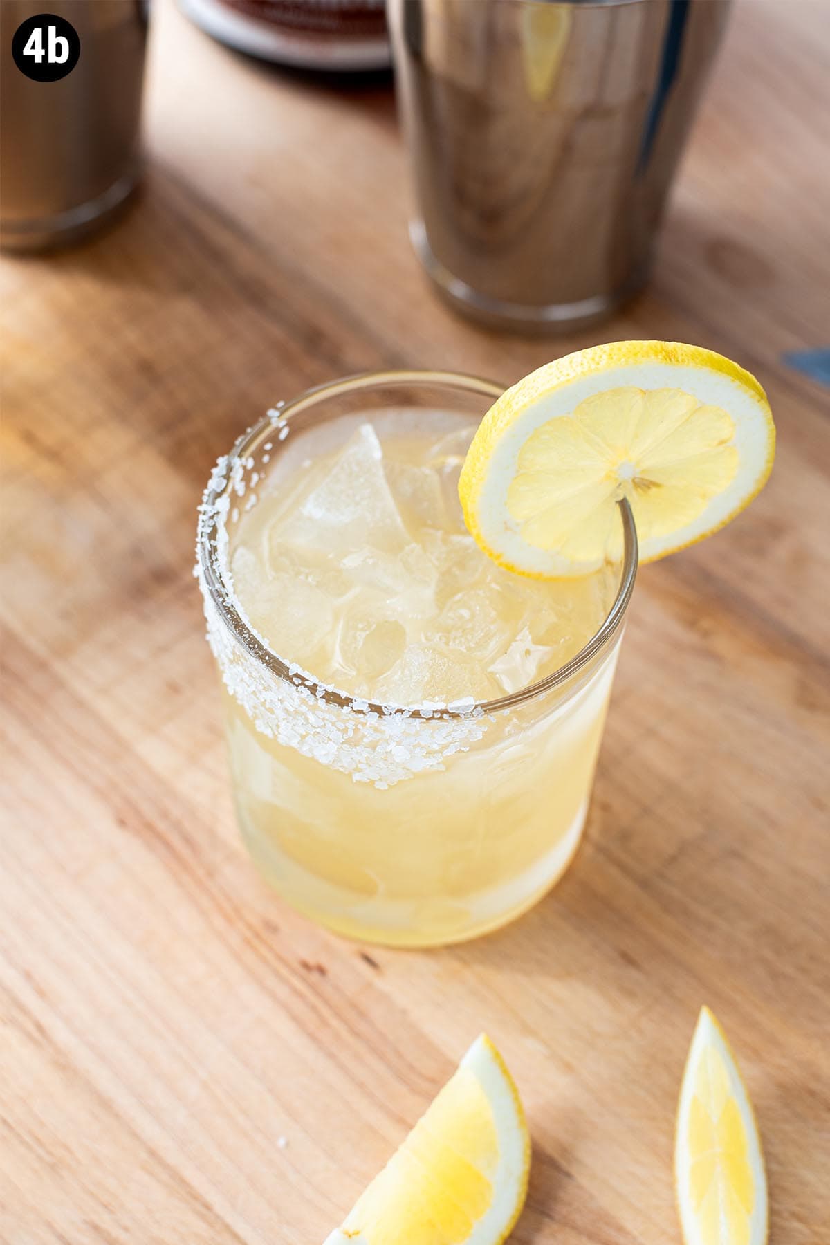 A golden margarita made with Grand Marnier with a salted rim and lemon wheel garnish sitting on a wooden cutting board.