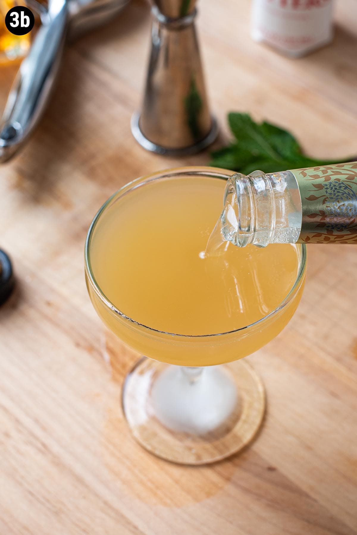 Topping a straight up bourbon cocktail with ginger beer in a chilled coupe glass.