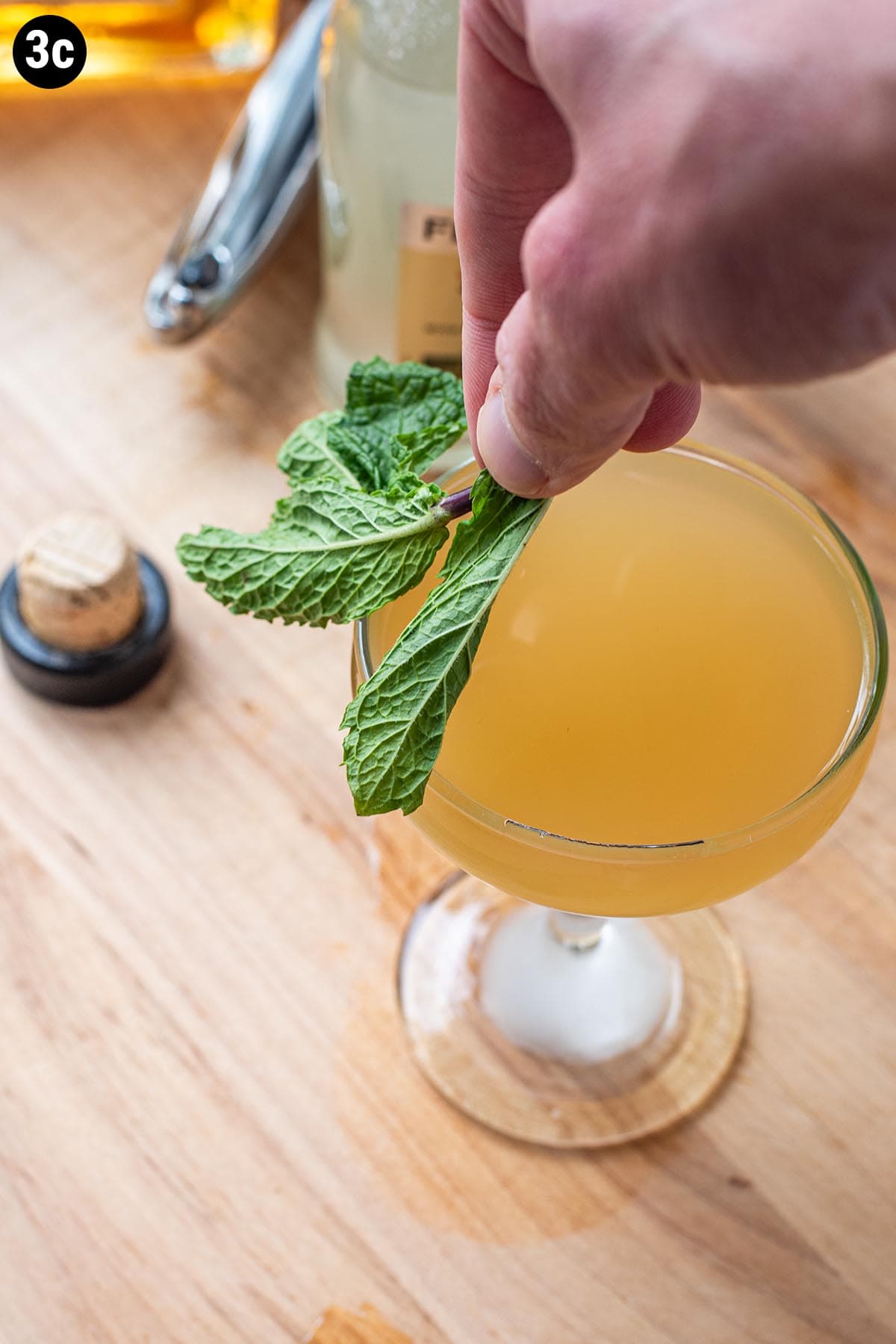 Wiping the rim of a bourbon cocktail in a chilled coupe glass with a sprig of fresh mint leaves.