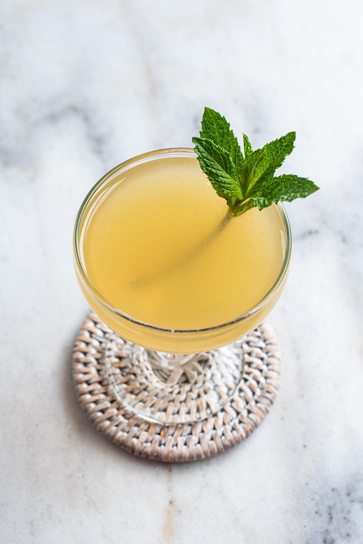 A golden bourbon cocktail served straight up in a small coupe glass and garnished with a small sprig of fresh mint.