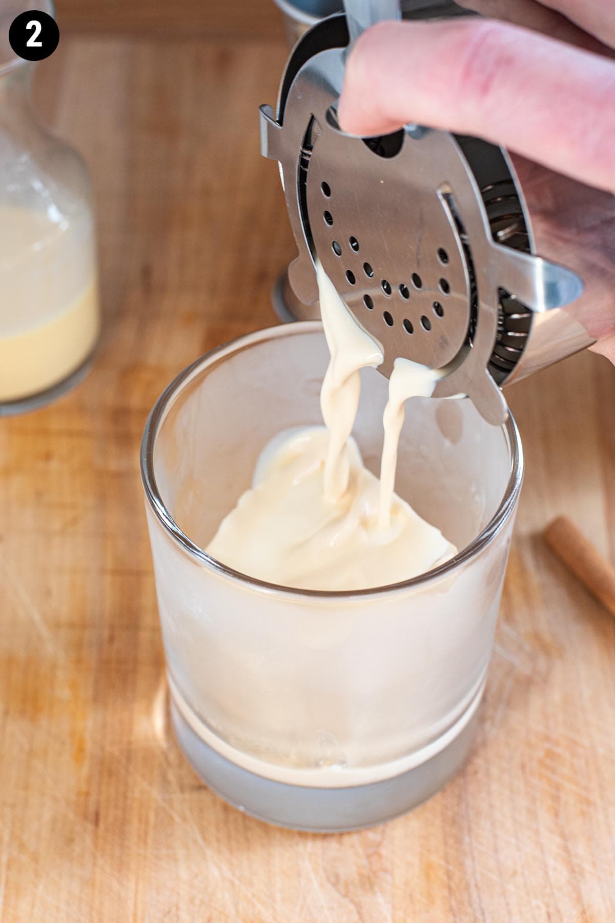 Straining an eggnog cocktail into a chilled double old fashioned glass with a large ice cube.