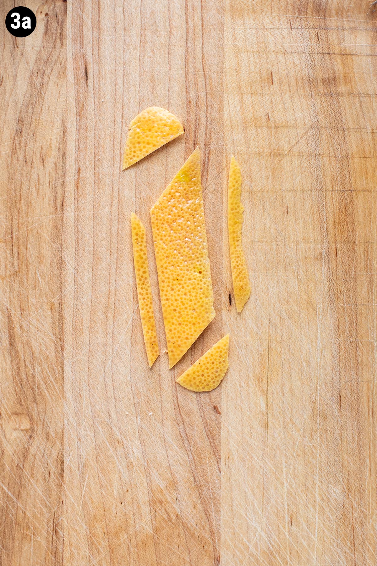 A grapefruit peel on a wooden cutting board sliced into a trapezoid shaped cocktail garnish.