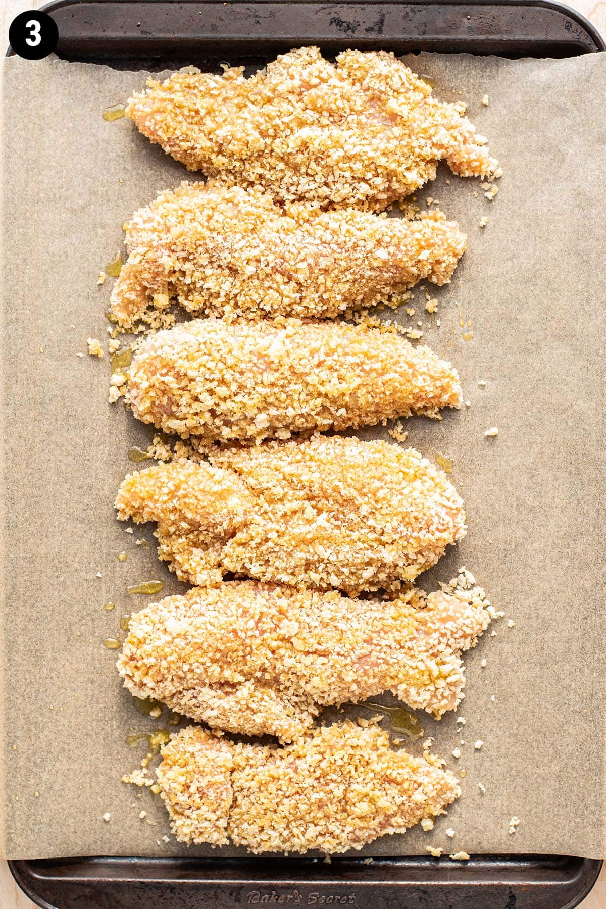 Unbaked panko crusted chicken tenders arranged in a row on a parchment-lined baking sheet.
