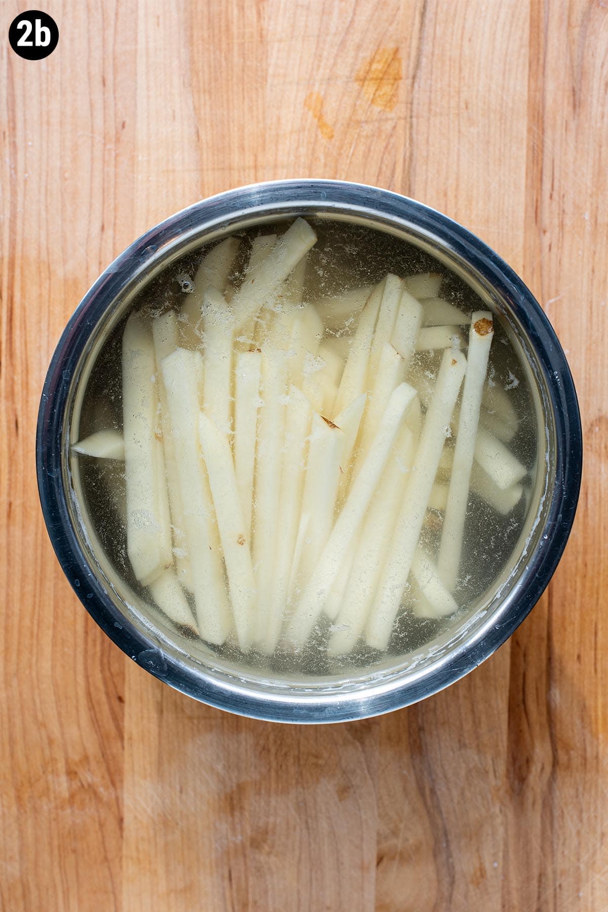 Hand cut uncooked French fries soaking in a stainless steel bowl with cool water.