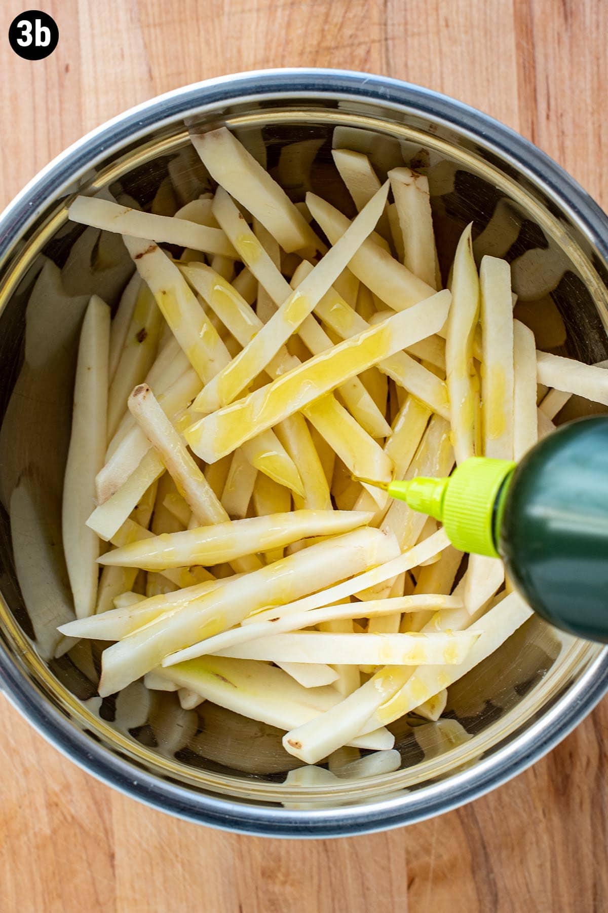 Olive oil being drizzle over uncooked hand cut French fries in a stainless steel bowl.