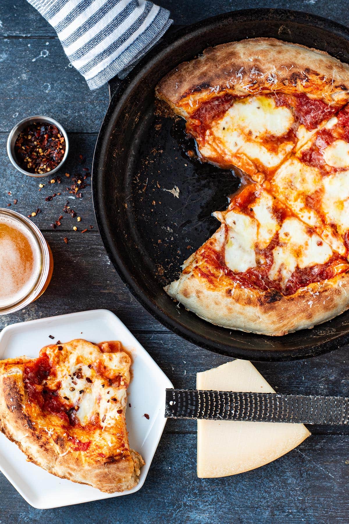 A cheesy golden Margherita pizza in a cast iron skillet next to a plate with a slice of pizza on it.
